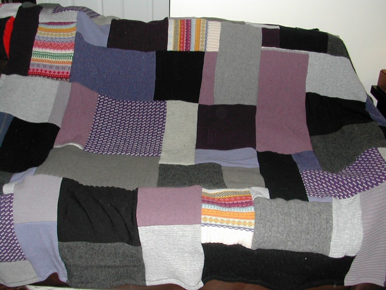 May include: A patchwork quilt made from different colored knitted fabrics. The quilt is made up of squares and rectangles of various sizes and colors, including gray, purple, black, and white.