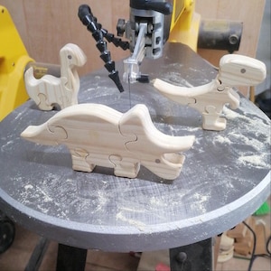 May include: Three unfinished wooden dinosaur puzzles are arranged on a grey surface near a band saw. The puzzles depict a Triceratops, a Tyrannosaurus Rex, and a Brontosaurus. The puzzles are made of light-coloured wood and are in various stages of completion.