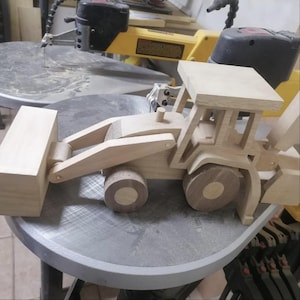 Puede incluir: Un juguete de madera hecho a mano con forma de excavadora. El juguete presenta una construcción de madera de color claro con un cucharón delantero, ruedas y una cabina. El diseño muestra la veta natural de la madera y está sobre una superficie circular.
