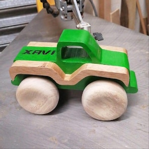Puede incluir: Un camión de juguete de madera con cabina verde y plataforma de madera natural. El camión tiene ruedas grandes, redondas y sin terminar. La palabra "XAVI" está impresa en negro en la plataforma.
