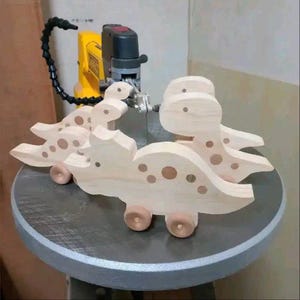 May include: Two wooden toy dinosaurs with brown spots and wheels. The dinosaurs are light-coloured and appear to be handmade. They are positioned on a grey surface, with a yellow and black tool in the background.