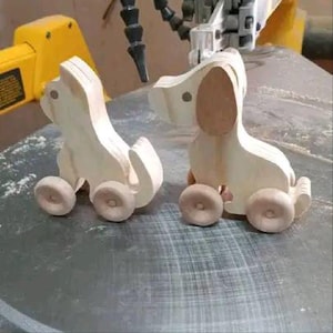 Op de afbeelding: Twee onafgewerkte houten hondenspeelgoed met wielen. De honden zijn lichtgekleurd hout, met afgeronde kenmerken en eenvoudige ontwerpen. De wielen zijn een lichtere tint hout. Het speelgoed staat op een grijze ondergrond.
