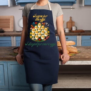 May include: A navy blue apron with a white strap and a colorful graphic of eggs with faces and the text "I came for the eggs. I stayed for more eggs."