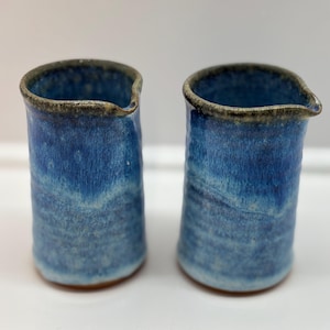 May include: Two handmade ceramic jugs with a blue glaze. The jugs have a small spout and a dark rim. The blue glaze has a gradient effect, transitioning from a darker shade at the top to a lighter shade at the bottom.