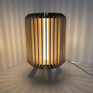 May include: A cylindrical wooden lamp with vertical slats, emitting a warm glow. The lamp sits on three white legs, casting a patterned shadow. A white cord extends from the base.