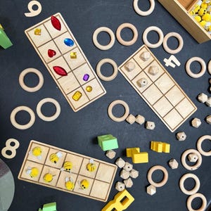 May include: Wooden educational toys, including number tiles, geometric shapes, and counting boards with colourful gems and bee designs. Also includes wooden rings and geometric blocks. The toys are arranged on a dark surface.