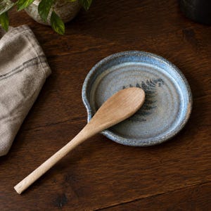 May include: A round, blue-glazed ceramic spoon rest featuring a dark fern leaf imprint at the centre. A light-coloured wooden spoon with a smooth, tapered handle rests diagonally across the surface of the ceramic dish.