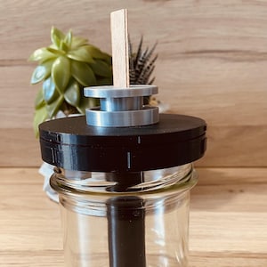 May include: A black and silver diffuser with a wooden stick inserted into the top. The diffuser is sitting on a wooden surface.