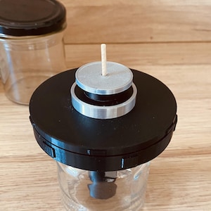 Puede incluir: Una tapa de frasco negra y plateada con un palo de madera que sobresale de la parte superior. La tapa está encima de un frasco de vidrio transparente.