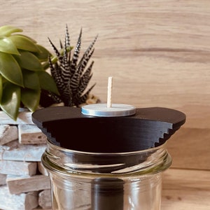 Puede incluir: Un portavelas negro con una pequeña vela redonda plateada en la parte superior. El portavelas se coloca encima de un tarro de vidrio transparente.
