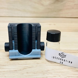 May include: A 3D-printed black and grey device with a cylindrical aperture, next to two small glass vials with black lids. The vials have white labels with the text "IDEE CREATION 3D". The items are on a wooden surface.