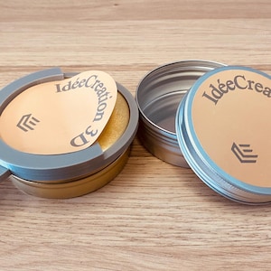 Peut inclure: Deux boîtes rondes en métal avec des couvercles dorés portant le texte IdéeCreation 3D et un logo. Une boîte est fermée, tandis que l'autre est partiellement recouverte par un cadre circulaire en plastique gris, révélant un intérieur doré.