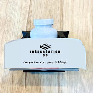 Op de afbeelding: Een witte fles met een witte dop staat op een wit en zwart 3D-geprint displaystandaard. De standaard heeft de tekst "IDEE CREATION 3D" en "Imprimez vos idées!" in het zwart.