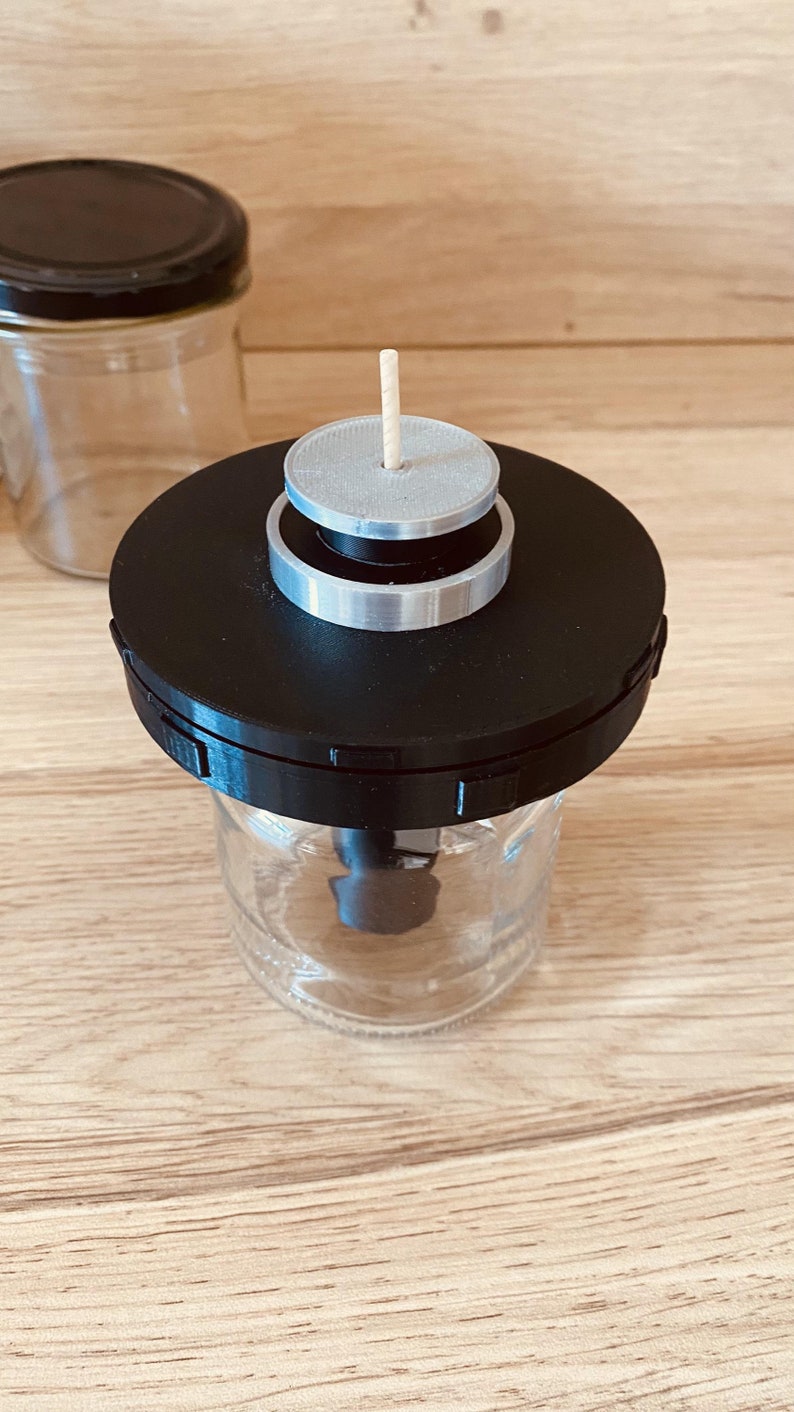 May include: A black and silver jar lid with a wooden stick sticking out of the top. The lid is on top of a clear glass jar.