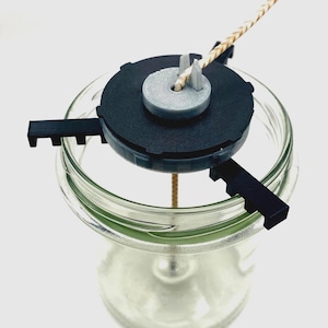 May include: A clear glass jar with a black lid and a beige wick. The lid has a circular design with a grey centre and three black tabs extending from the sides. A beige wick extends from the centre of the lid.