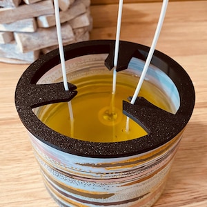 Puede incluir: Un tarro de vidrio para velas con tapa negra y un diseño abstracto colorido. El tarro está lleno de cera amarilla y tiene tres mechas blancas.