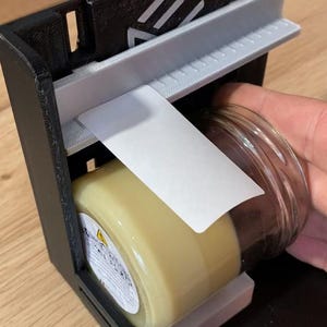 Peut inclure: Un distributeur d'étiquettes imprimé en 3D noir et gris avec une étiquette blanche en cours de distribution. Le distributeur d'étiquettes a un bouton sur le côté et un pot en verre transparent avec une étiquette blanche dessus.