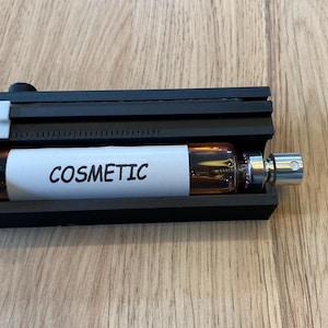 Könnte beinhalten: Ein schwarzer rechteckiger Koffer, der eine kleine Glasflasche mit silbernem Verschluss enthält. Die Flasche enthält eine braune Flüssigkeit und hat ein weißes Etikett mit dem Wort "COSMETIC". Der Koffer steht auf einer Holzoberfläche.
