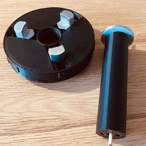 May include: A black 3D printed spool with three gray tabs and a black 3D printed cylinder with a silver pin at the bottom.