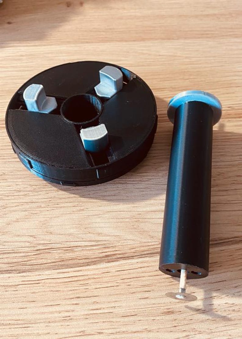May include: A black 3D printed spool with three gray tabs and a black 3D printed cylinder with a silver pin at the bottom.