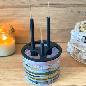 May include: A black candle holder with a colorful abstract design. The holder has three slots for candles and is made of wood. The candles are white and have black wicks.