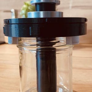 May include: A black and silver 3D printed device with a wooden dowel attached to the top. The device is sitting on top of a clear glass jar.