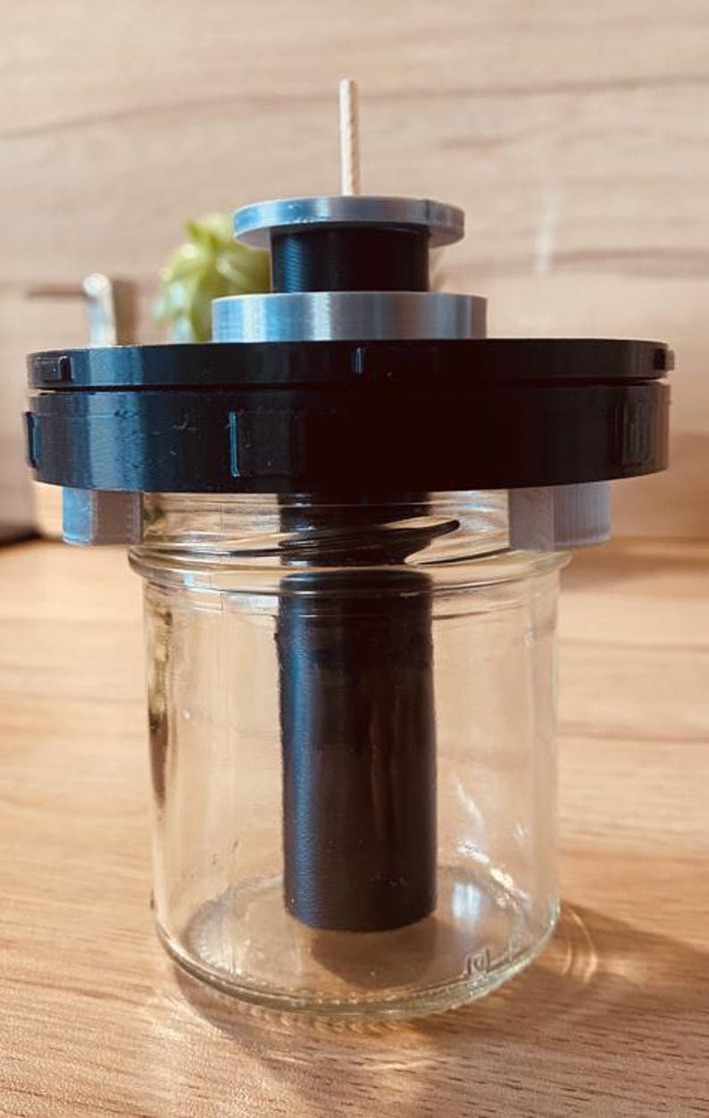 May include: A black and silver 3D printed device with a wooden dowel attached to the top. The device is sitting on top of a clear glass jar.