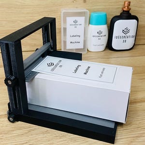 Può includere: Una macchina etichettatrice nera con una scatola bianca e un'etichetta stampata è mostrata. L'etichetta recita "INTEGRATION 3D" e "Labeling Machine". Altri elementi includono una bottiglia bianca e una bottiglia nera.