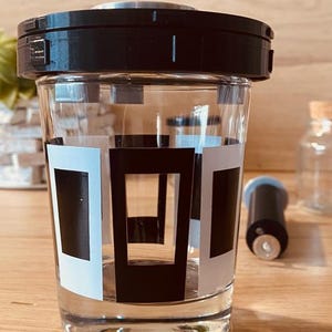 May include: A clear glass jar with a black lid and a black and white geometric design. The jar is filled with a clear liquid.