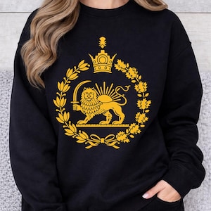 Puede incluir: Sudadera negra con un emblema dorado de un león sosteniendo una espada, rodeado por una corona y una corona de laurel. El diseño está centrado en la parte delantera. La sudadera es de cuello redondo.