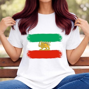 Puede incluir: Camiseta blanca con el diseño de la bandera iraní. El diseño incluye una franja verde, una franja roja y un centro blanco con un emblema de león y sol dorado. La camiseta es de manga corta.