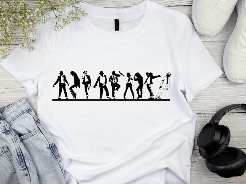 May include: White t-shirt featuring a black silhouette of a group of people in various dance poses. The design is a line of figures in motion, with a focus on dance. The shirt is displayed on a wooden surface, with jeans, headphones, and shoes in the background.