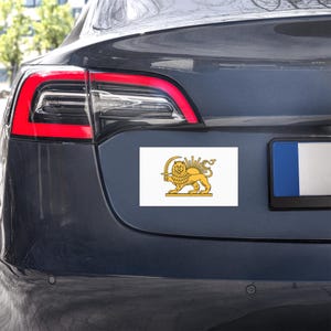 Puede incluir: Un imán rectangular blanco para coche con un león dorado sosteniendo una espada, con un diseño de sol radiante encima. El imán está fijado en la parte trasera de un coche gris oscuro.