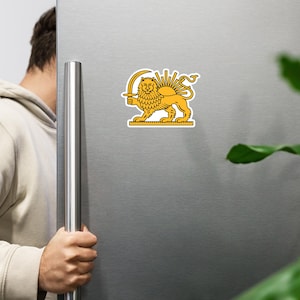 Puede incluir: Una pegatina amarilla y negra con un león sosteniendo una espada, con un sol detrás. La pegatina está adherida a la puerta plateada de una nevera. El león está de pie.