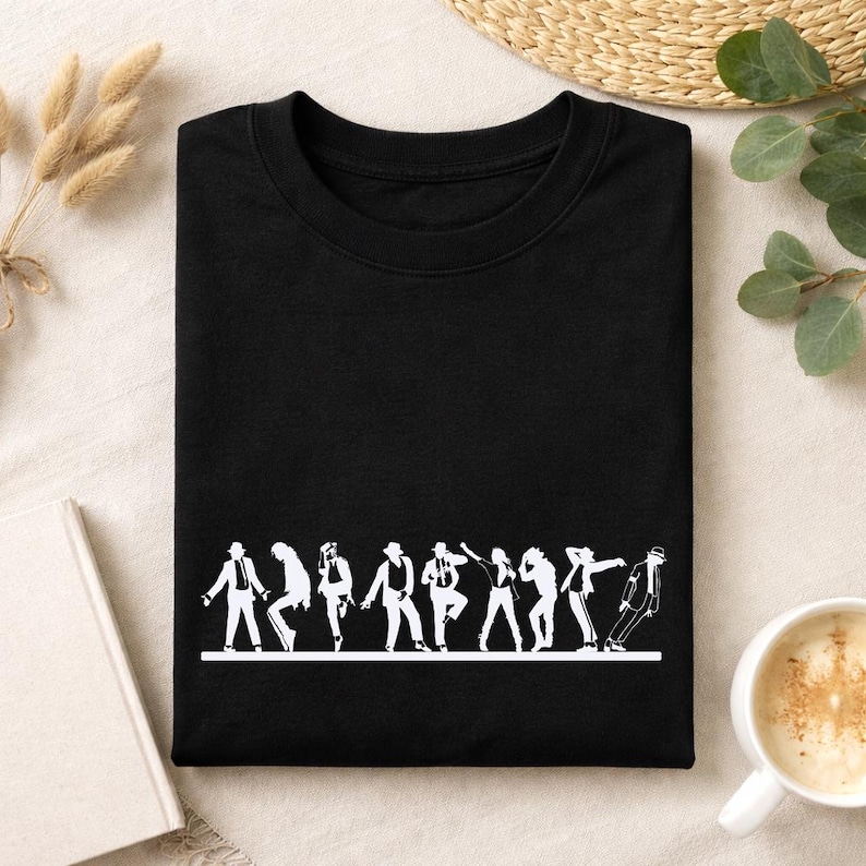 May include: Black crew neck t-shirt with a white graphic of a line of dancers in various poses. The dancers are in silhouette, and the design is printed on the front of the shirt. The shirt is folded neatly.