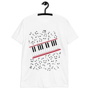 Pode incluir: Camiseta branca com um desenho de teclado de piano. O teclado tem teclas pretas e brancas e uma borda vermelha. Notas musicais pretas estão espalhadas ao redor do teclado. A camiseta está pendurada em um cabide preto.