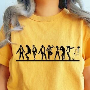 May include: Mustard yellow t-shirt featuring a black silhouette of a group of people in various dance poses. The graphic is a line of figures, each in a different dance move, on a horizontal black line.