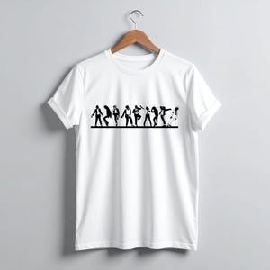 May include: White t-shirt with a black graphic of stylized figures in various dance poses. The design spans the chest area of the shirt. The shirt is hanging on a wooden hanger against a plain gray background.