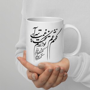 Könnte beinhalten: Weiße Keramiktasse mit schwarzer Kalligraphie. Die Tasse hat einen gebogenen Henkel und eine glänzende Oberfläche. Die Schrift scheint eine künstlerische Darstellung eines Zitats oder Gedichts zu sein. Die Tasse wird von einer Person gehalten.