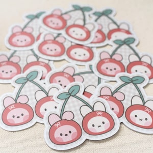 May include: A collection of stickers featuring a cartoon cherry design. Each sticker has a red cherry with a cute face, a white bow, and green leaves. The background is a light beige color.