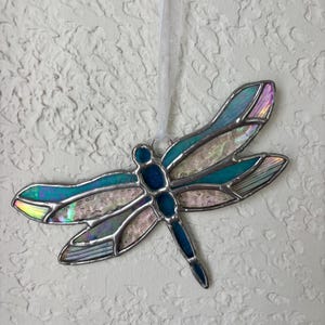 May include: A stained glass dragonfly ornament featuring iridescent teal and pearlescent white wings with dark metal soldering. The dragonfly has a segmented blue body and is suspended by a thin white ribbon for hanging display purposes.