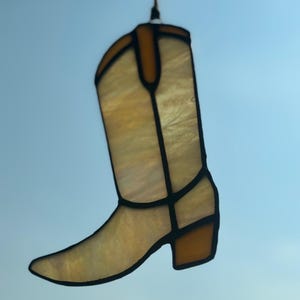 May include: A stained glass cowboy boot with a light amber and brown color scheme. The boot is outlined in black and hangs from a black wire. The boot has a pointed toe and a block heel.