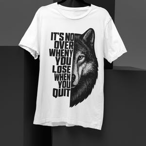 Peut inclure: T-shirt blanc avec une illustration en noir et blanc d'un loup sur le côté droit. Le côté gauche affiche le texte "IT'S NO OVER WHEN YOU LOSE WHEN YOU QUIT" verticalement. Design simple et audacieux.
