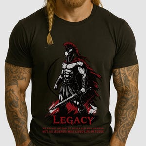Puede incluir: Camiseta gris oscuro con un guerrero espartano con capa y casco rojos, sosteniendo una espada. La palabra "LEGACY" está impresa debajo, junto con una cita sobre leyendas. El diseño es en negro, blanco y rojo.