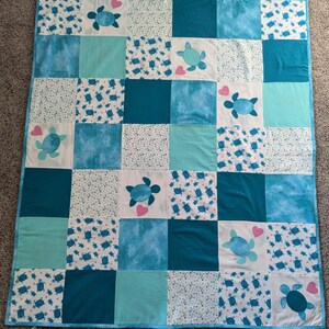 May include: A patchwork quilt featuring a sea turtle theme. The quilt is composed of squares in shades of blue and white, with sea turtle appliqués and pink heart accents. The quilt is bordered with a light blue trim.