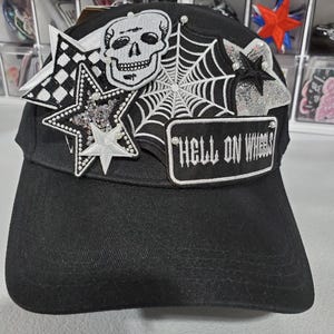 May include: Black baseball cap with embroidered patches. Patches include a skull, spiderweb, stars, and the text "HELL ON WHEELS". The cap has a curved brim and is decorated with small pearl-like beads.
