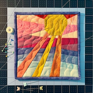 Può includere: Un piccolo arazzo quadrato trapuntato con un disegno a raggiera. Il sole è giallo, con raggi arancioni, rosa e viola. Lo sfondo è un gradiente di rosa, viola e blu. Il pezzo è incorniciato con un bordo blu.