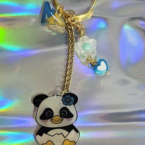 K&ouml;nnte beinhalten: Ein goldfarbener Schl&uuml;sselanh&auml;nger mit einem Panda-B&auml;ren-Anh&auml;nger, einem blau-gelben Berg-Anh&auml;nger und einer Kette mit Blumen- und Herz-Anh&auml;ngern. Der Panda ist schwarz, wei&szlig; und gelb. Das Wort "Lunar" steht unten.