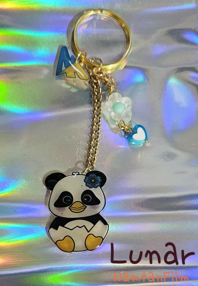 K&ouml;nnte beinhalten: Ein goldfarbener Schl&uuml;sselanh&auml;nger mit einem Panda-B&auml;ren-Anh&auml;nger, einem blau-gelben Berg-Anh&auml;nger und einer Kette mit Blumen- und Herz-Anh&auml;ngern. Der Panda ist schwarz, wei&szlig; und gelb. Das Wort "Lunar" steht unten.