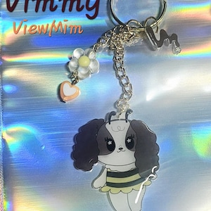 May include: A keychain featuring a cartoon dog character dressed as a bee with black and yellow stripes, attached to a silver ring. It includes a small charm of a clear flower with a yellow center, a peach heart bead, and a silver letter M charm.